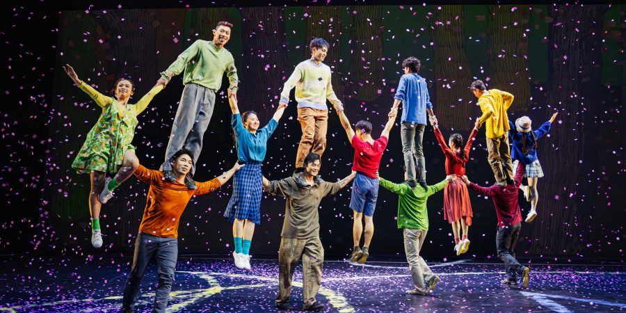 A crew of performers as part of a circus, dressed colourfully, hold hands while balanced atop each other.